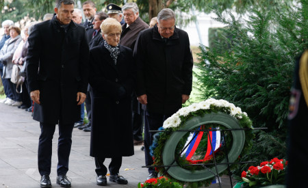 Skupina ljudi se poklanja pri Grobnici narodnih herojev. Moški položijo venec z belimi rožami in trakom v nacionalnih barvah.