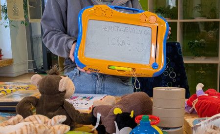 ženska drži v rokah tablo piši-briši, pred njo na mizi so igrače